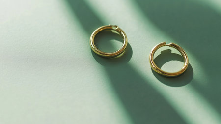 A pair of elegant gold hoop earrings resting on a soft green surface, illuminated by natural light and creating striking shadows that enhance their beauty.の素材