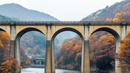A tranquil autumn scene featuring a bridge arching gracefully over a calm river, framed by colorful foliage and majestic mountains in the background.の素材