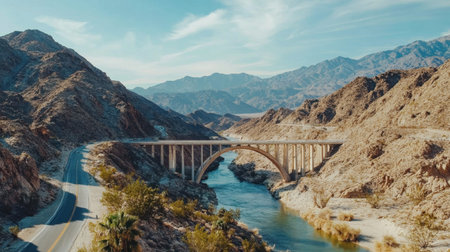 This stunning image captures a picturesque desert bridge arching over a clear river, framed by majestic mountains under a bright blue sky. Perfect for nature lovers.の素材