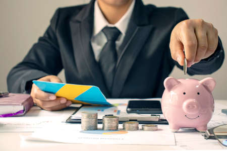 A businessman holds a coin on the piggy bank to save money for investment ideas, savings and sustainable investments.の写真素材