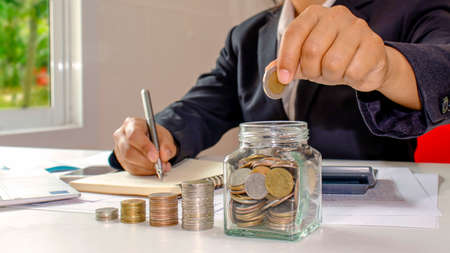 Business women who are putting coins in a jar of money, including a notebook on a wooden desk for work, ideas for saving money and managing accounts.の写真素材