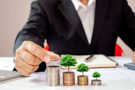 The hands of businessmen are planting trees growing on coins, including notebooks on wooden desks for work, ideas for saving money and managing accounts.の写真素材