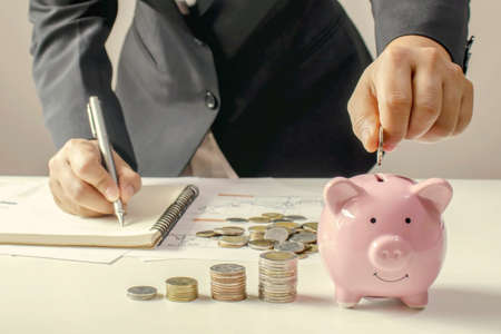Close-up of a woman holding a coin in a pig piggy bank and recording income, expenses, ideas for calculating expenses and saving money.の写真素材