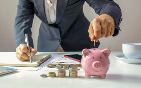 Close-up of a woman holding a coin in a pig piggy bank and recording income, expenses, ideas for calculating expenses and saving money.の写真素材