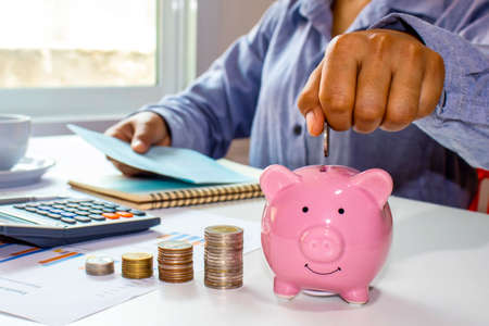 Close-up of a woman holding a coin in a pig piggy bank and recording income, expenses, ideas for calculating expenses and saving money.の写真素材