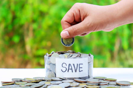 Hand holding a coin in a piggy bank on a natural green background, concept to grow and save money.の写真素材