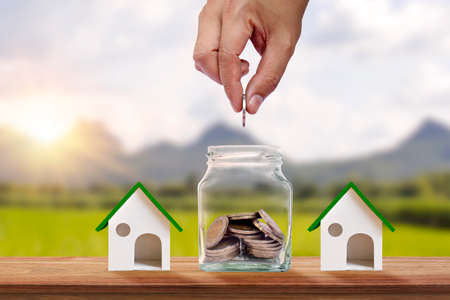 Hand holding a coin in a piggy bank and house model on the wooden floor, financial concept.の写真素材