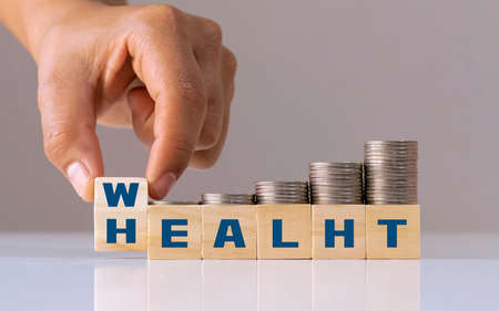 Hand flips a wooden cube with the words Wealth for Health and a growing pile of coins. Life insurance investment ideas and health care concepts.の写真素材