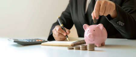 A businessman puts a coin in a piggy bank and takes notes on finances. Concept of saving money and checking accounting documents.の写真素材