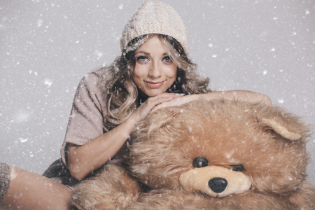 Casual smiling young woman in knitted clothes holding big soft teddy bear on snowy background. Studio shotの写真素材