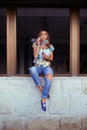 Young woman portrait blowing soap bubbles. Sitting in the window frameの写真素材