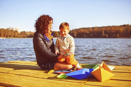Mom and son playing with paper boats by the lake. Warm filter and film effectの写真素材