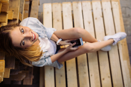 Portrait of charming girl sitting on a pallet using smart phoneの写真素材