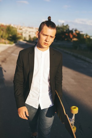 Vintage image of a man with longboard going on road,hipster young teen boy, sunset lights,mans hairstyle, mans fashion, urban street styleの写真素材