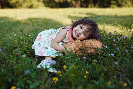 Cute smiling baby girl hugging soft bear toy, sweet kid having fun outdoors in day care, laying down on green grass in the parkの写真素材