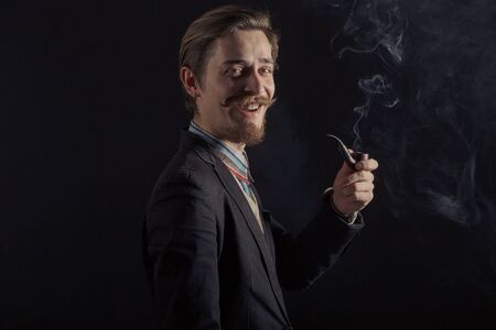 handsome bearded stylish business man in elegant black suit posing in studio on black background, smoking pipeの写真素材