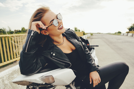 Biker girl in a leather clothes on a white motorcycle. Sexy young woman biker sitting on her motorcycle on a road. Adventure concept.の写真素材