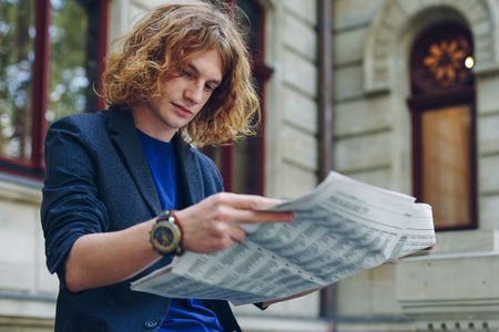 Portrait young reddish, red haired attractive man with curly hair, reading a newspaper sitting leg over leg near an urban old style building. Youth in action leader getting to know world, city news.の写真素材