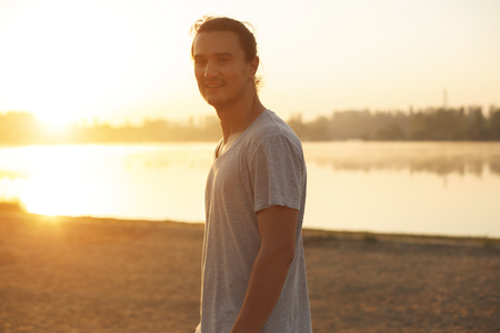 Handsome man smiling showing teeth in the park, on the lake while looking at sunrise. Portrait of athletic capoeira man on city beach background, wearing gray t-shirt, resting after training.の写真素材
