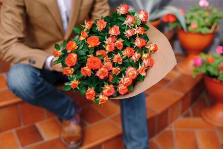 Focus on bunch of roses, folded in craft paper holded by man talking on phone sitting outdoors, wearing jeans, wool jacket. Flower delivery. Valentines day. Love is in the air. Postcard flowers.の写真素材
