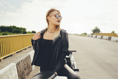 Attractive young woman posing on a motorbike wearing fashionable sunglasses, leather jacket, stylish haircut. Confident face expression, hairstyle, sensual biker femaleの写真素材