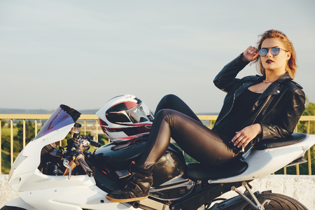 Beautiful woman biker laying on a bike relaxing, posing, smiling, looking to camera, enjoying the moment wearing sunglasses and leather clothes with road and sky in background.の写真素材