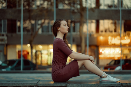 Side view of pretty young woman with fairytale face features enjoy city view with lights in the background. Beautiful urban girl portrait wearing dress and white sneakers.の写真素材
