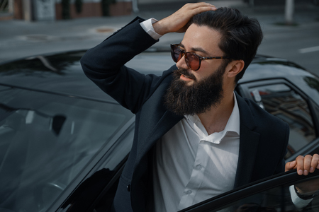 Beautiful, stylish, bearded man, smiling, happy while standing near car touching his brunette hair wearing fashionable sunglasses. Portrait attractive man. Life lover. City people.の写真素材