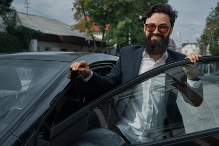 Beautiful, stylish, bearded man, smiling happy while standing near car, wearing a black jacket, white shirt and fashionable sunglasses. Portrait attractive man. Life lover. City people.の写真素材