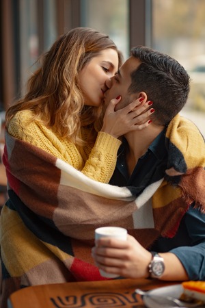 Young couple embracing and kissing covered with a warm plaid blanket and a cup of coffee with windows in the backgroundの写真素材