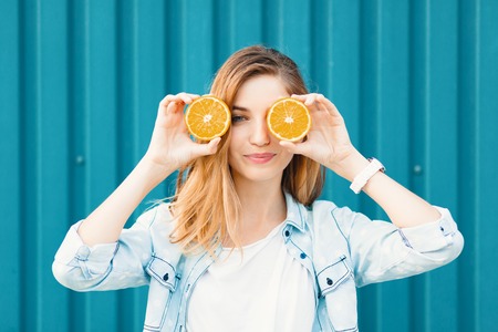 Carefree young beautiful girl using two halfs of oranges instead of glasses over her eyes over blue background.の写真素材