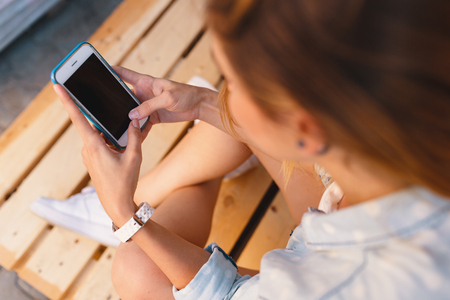Pretty woman holding the phone in hands while touching the screen and sitting on wood pallets, Cropped image top angle view.の写真素材