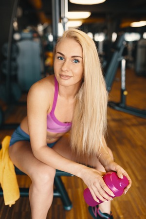 Pretty woman with long blond hair relaxing in the gym with a pink bottle of water with equipment in the background and beautiful wood floor.の写真素材