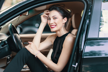 Gorgeous woman laughing, happy while sitting on the front seat in a black car with the opened door. Refined beautiful female with classic black clothes, red lips, shiny white teeth. Close-up.の写真素材