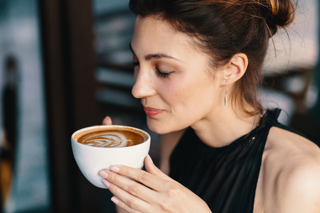 Coffee. Refined Woman Enjoying Cappuccino or Latte with Closed Eyes on a Vibrant Background Indoors. Cup of Hot Beverageの写真素材