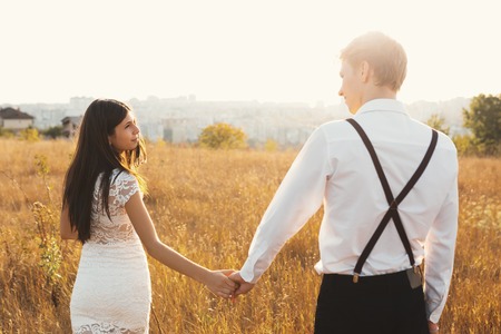 A rear view shot of a young woman walking with her boyfriend holding hands on grass field. A couple enjoying a walk through golden grassland with the city on the horizon on a sunny day.の写真素材