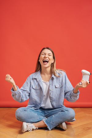 Smiling casual girl holding game joystick controller happily raise hands, celebrate winning game during a holiday on red background with copy space.の写真素材