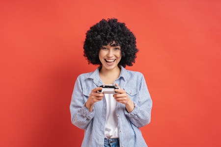 Excited delighted woman playing video games, while holding a game console in hands isolated on red background.の写真素材