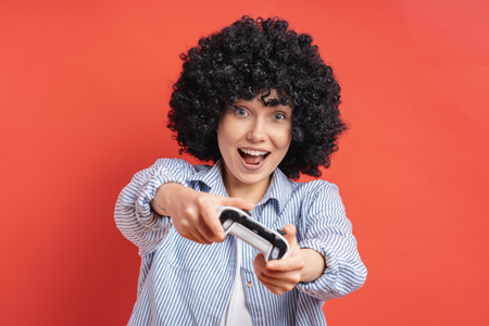 Excited delighted woman playing video games, while holding a game console in hands isolated on red...