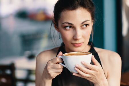 Coffee. Refined Woman Enjoying Cappuccino or Latte on a vibrant background indoors. Cup of Hot...