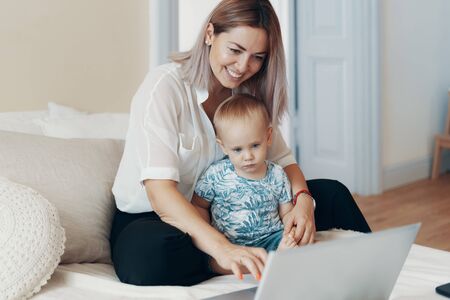 Young mother with her child working on laptop in bedroom at home. Multi-tasking, freelance and motherhood conceptの写真素材