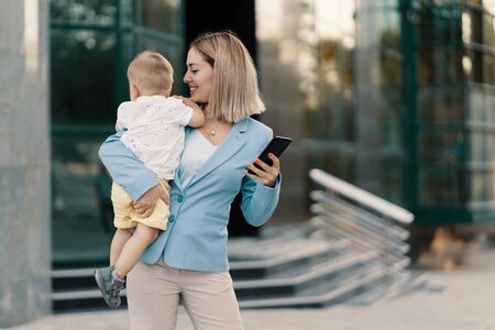 Successful business woman in blue suit with baby. Successful mother in business walking with baby in her hands and texting message on cellphoneの写真素材