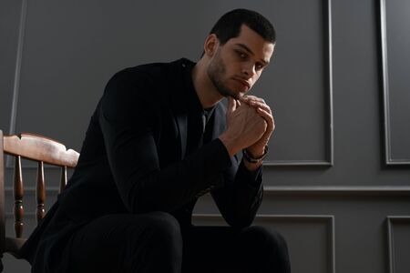 Young handsome businessman in black shirt and black suit. Confident young businessman posing on gray background sitting on chairの写真素材