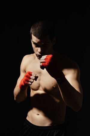 Boxing male portrait standing in a rack on black background with red bandages on his hands. Light and shadows play.の写真素材
