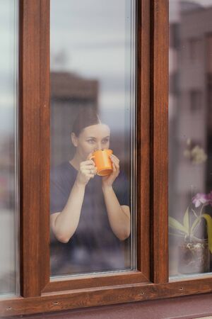 Rear view of beautiful woman looking out the window at the cityscape drinking tea while staying home.の写真素材