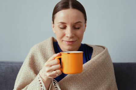 Happy woman recovered from a flu enjoying a cup of tea wrapped in a warm blanket at home on the sofa