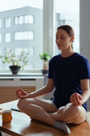 Young woman relaxing in yoga poses, sitting on a wood table in a modern interior with windows in the...