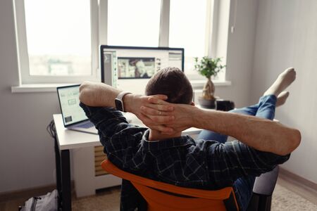 Man relaxing while working remotely from home holding legs on the table looking at the computer stretching hands behind his headの写真素材