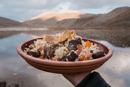 Man hand holding a clay plate with delicious food on lake and mountain background. Delicious rice...