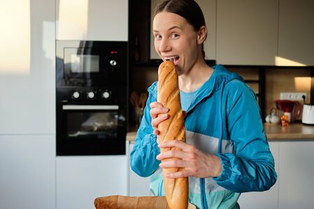 Cheerful young woman wants to bite a fresh and delicious baguette holding it with bough hands...